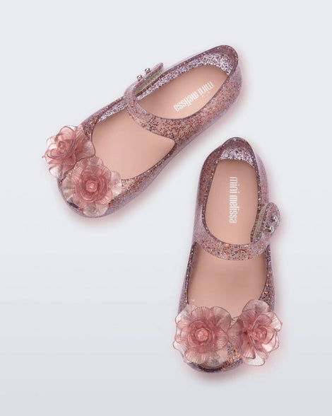 Top view of a pair of Mini Melissa Ultragirl ballet flats in Clear Glitter Pink colorway with 3D printed flower applique on the top of the toe. There is a single strap across the top of the foot with a double snap closure. Both shoes have 2 flower appliques on the top of the two