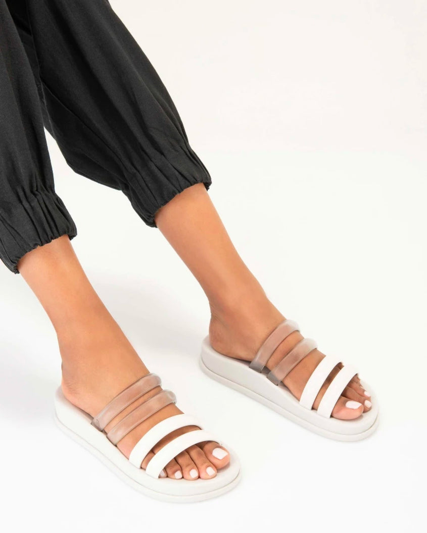 A model's legs in black pants, wearing a pair of white/clear Melissa Soft Wave Slides with 4 straps: two clear and two white.