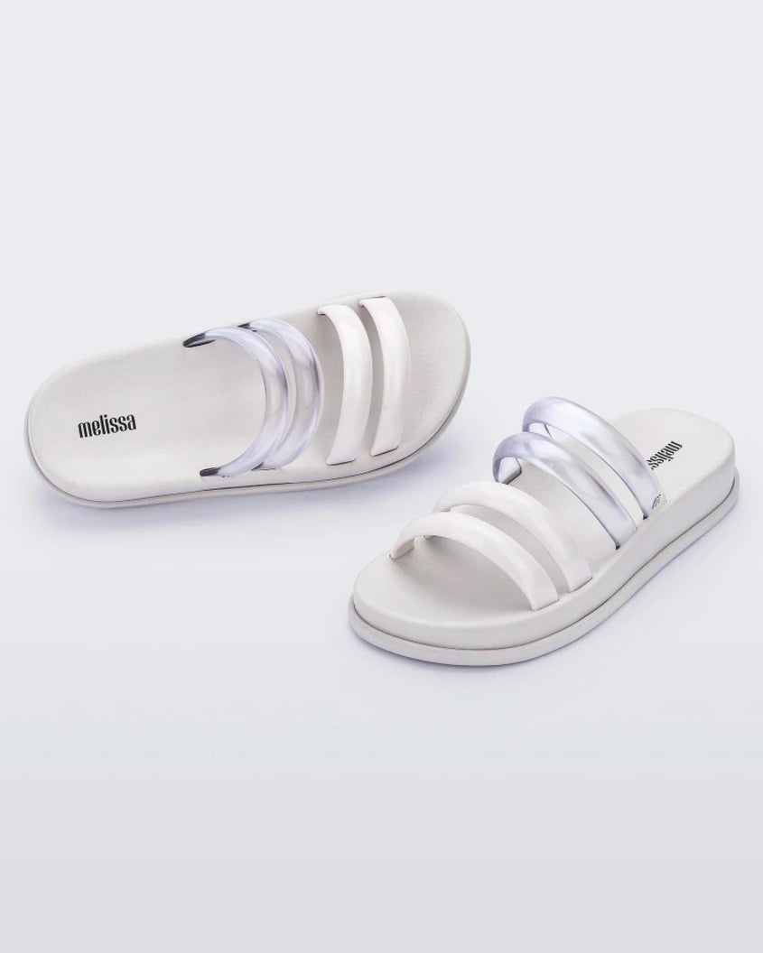 An angled front and top view of a pair of white/clear Melissa Soft Wave Slides with 4 straps: two clear and two white.