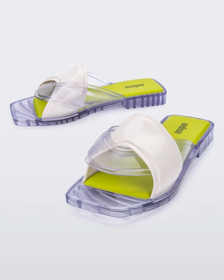 An angled front and side view of a pair of white/clear/yellow Melissa Brigitte slides with a white weft designed upper strap, clear lower strap and yellow insole.