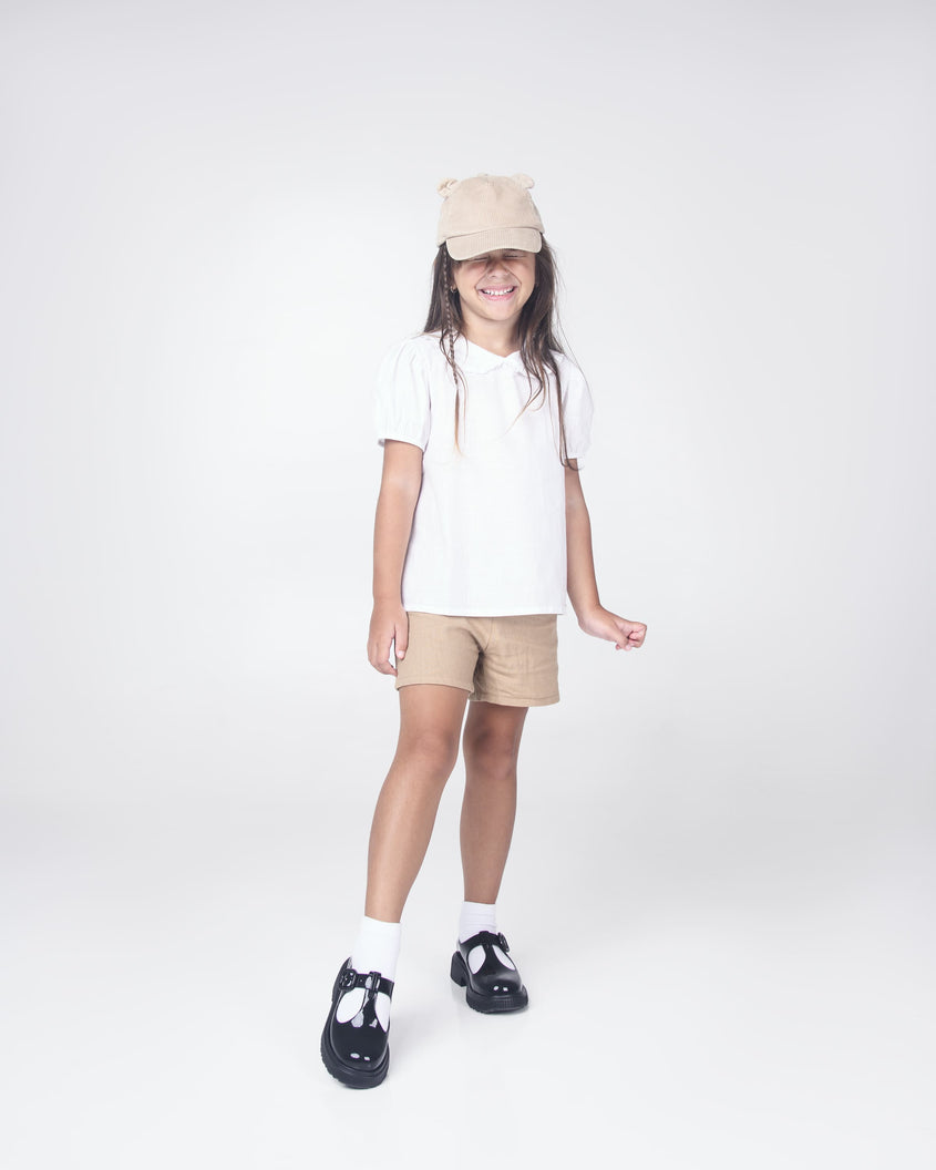 A model posing for a picture in a tan hat, white top, tan shorts, white socks and a pair of black Mini Melissa Jackie loafers with two cut outs and a buckle detail strap.