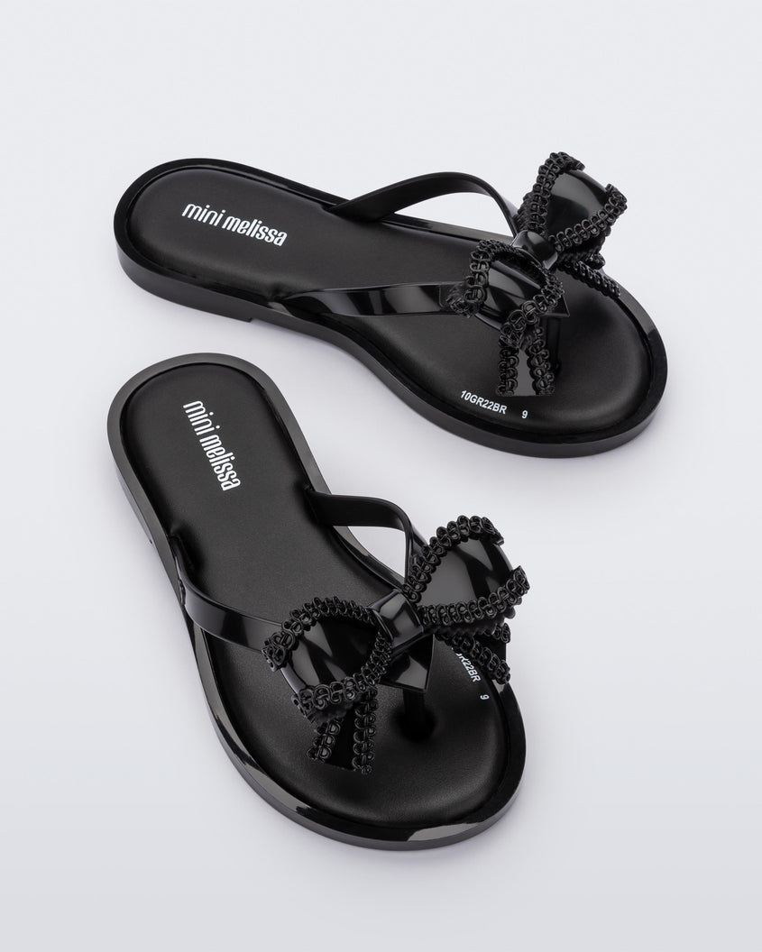 An angled front and side view of a pair of Black Mini Melissa Slim flip flops with a lace like bow detail on the front straps.