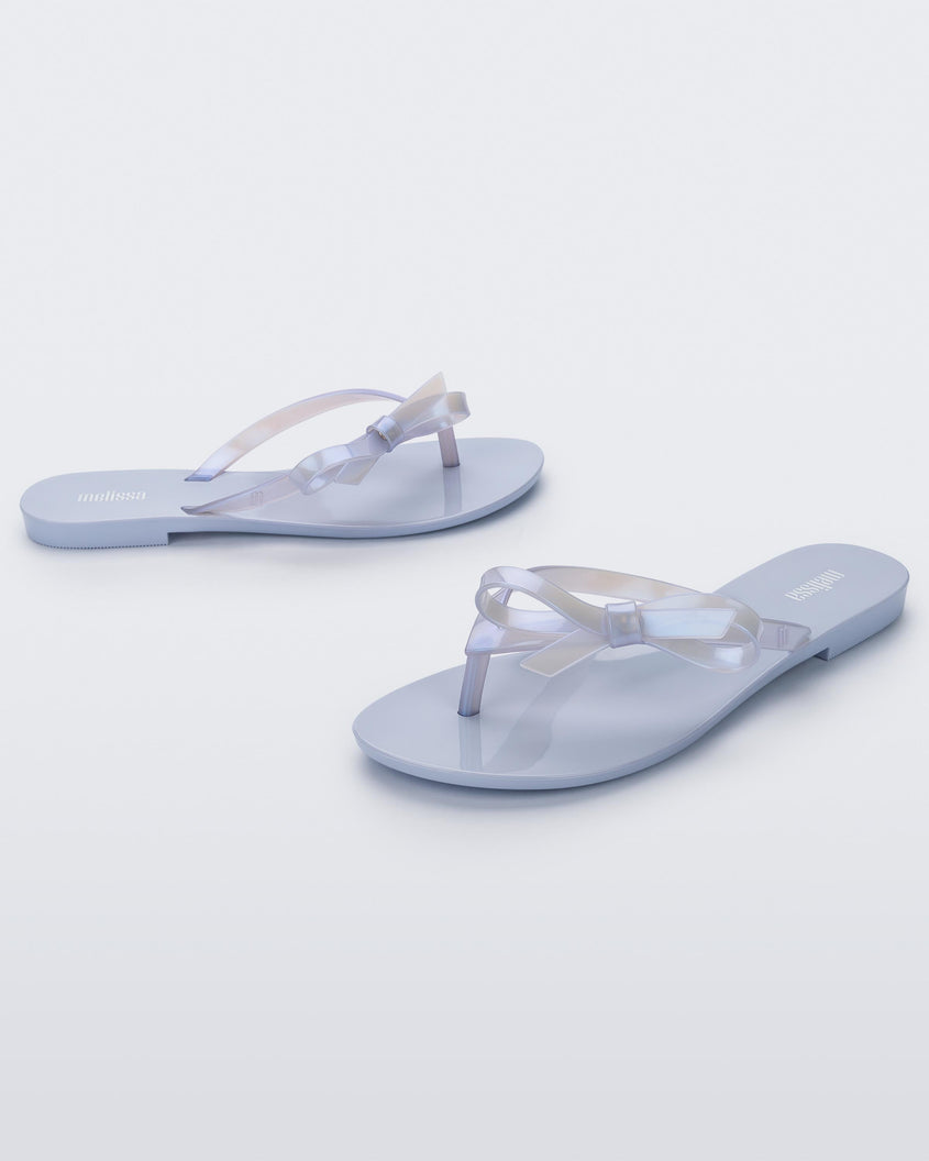 An angled front and side view of a pair of Lilac/Pearly blue Melissa Harmonic Sweet flip flops with a lilac/pearly blue sole, straps and bow on top.