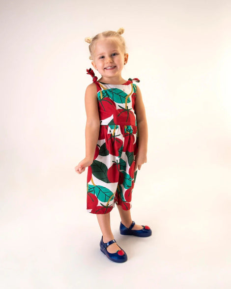 A young model posing for a picture in a white, red, and green patterned jumpsuit of a pair of Metallic Blue/Red Mini Melissa Sweet Love Princess flats with a metallic blue base, a top strap, Snow White in script on the side, an apple detail on the toe, and a drawing of Snow White on the sole.