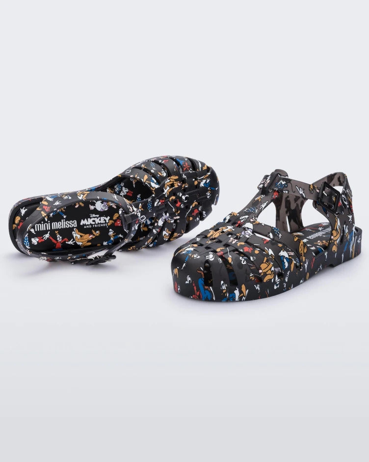 Angled view of a pair of Mini Melissa Possession fisherman sandals in black with all over Disney character print. There is a buckle closure on the side of the ankle strap. 