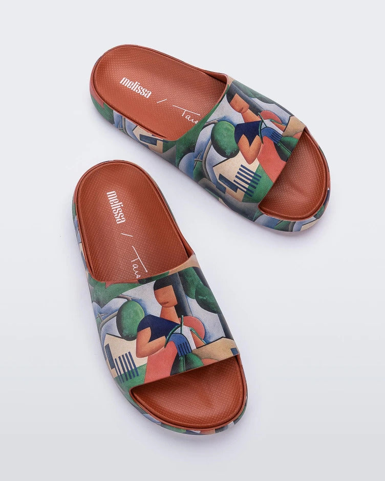 Top view of a pair of tan multicolor Melissa Cloud slides with an abstract painting design of a house, person, and farmland.