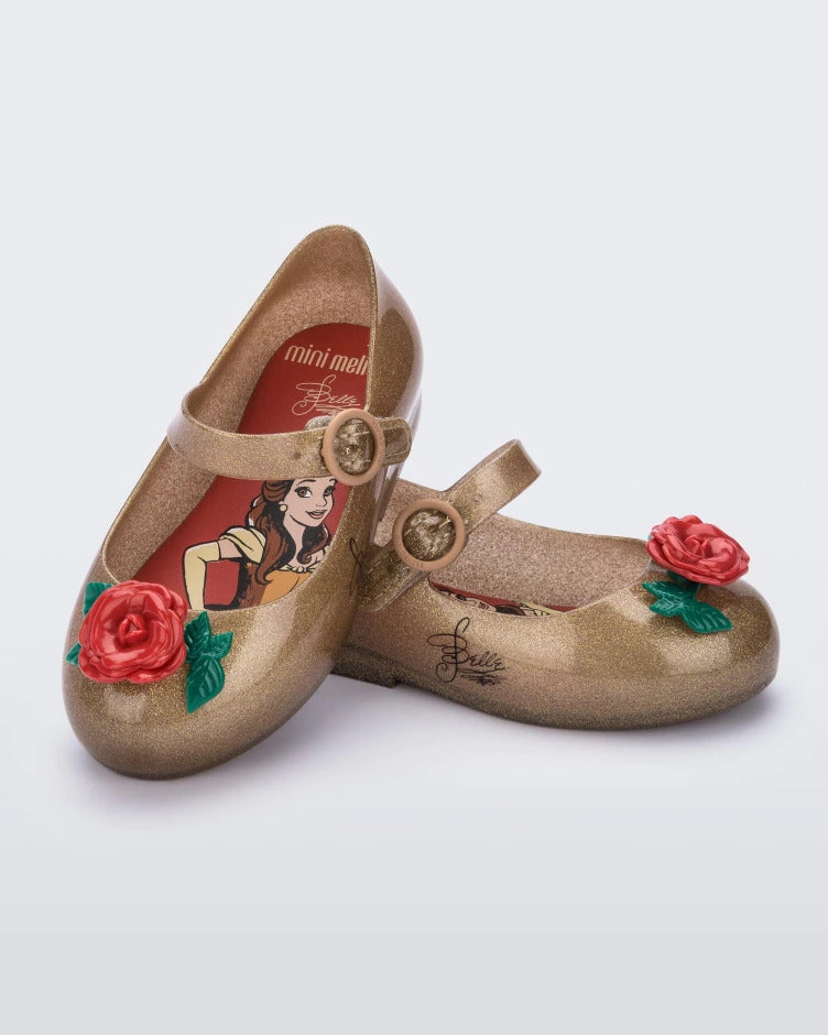 An angled top and side view of a pair of gold/red Mini Melissa Sweet Love Princess flats with a gold base with Belle in script on the side, a top strap, a rose detail on the toe and a drawing of Belle of Beauty and the Beast on the insole.