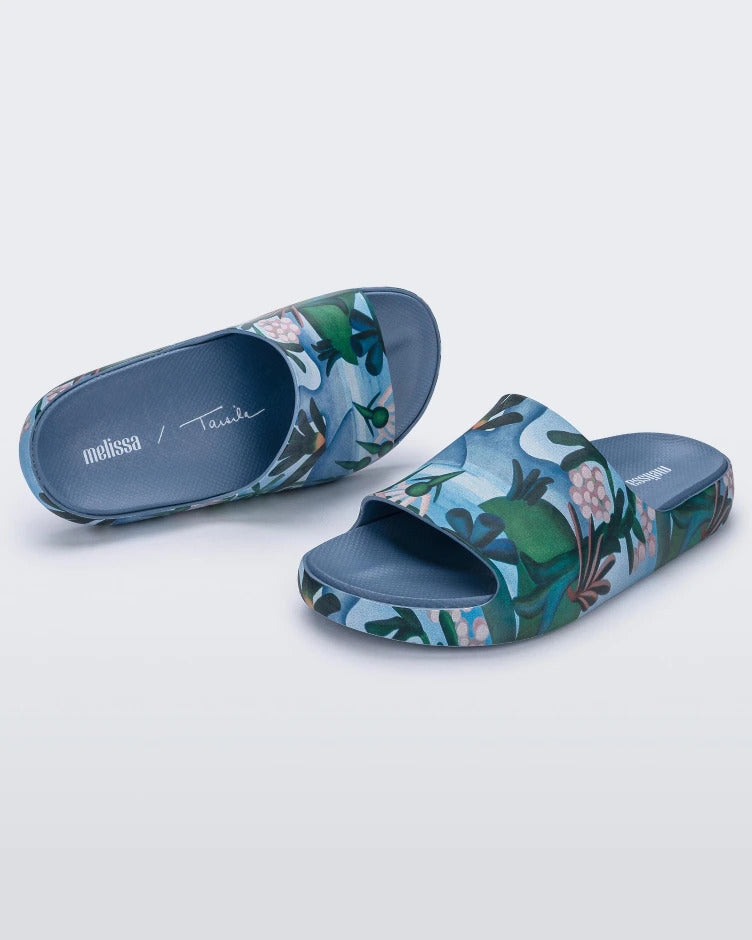An angled front and top view of blue multicolor Melissa Cloud slides with an abstract painting design of plants, water and lagoon.