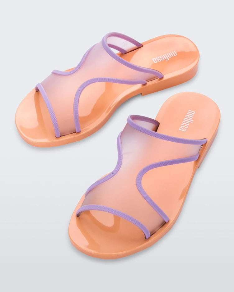 An angled top view of a pair of orange/lilac Melissa Bikini Slides with a two transparent purple straps conjoining in the middle and an orange insole.