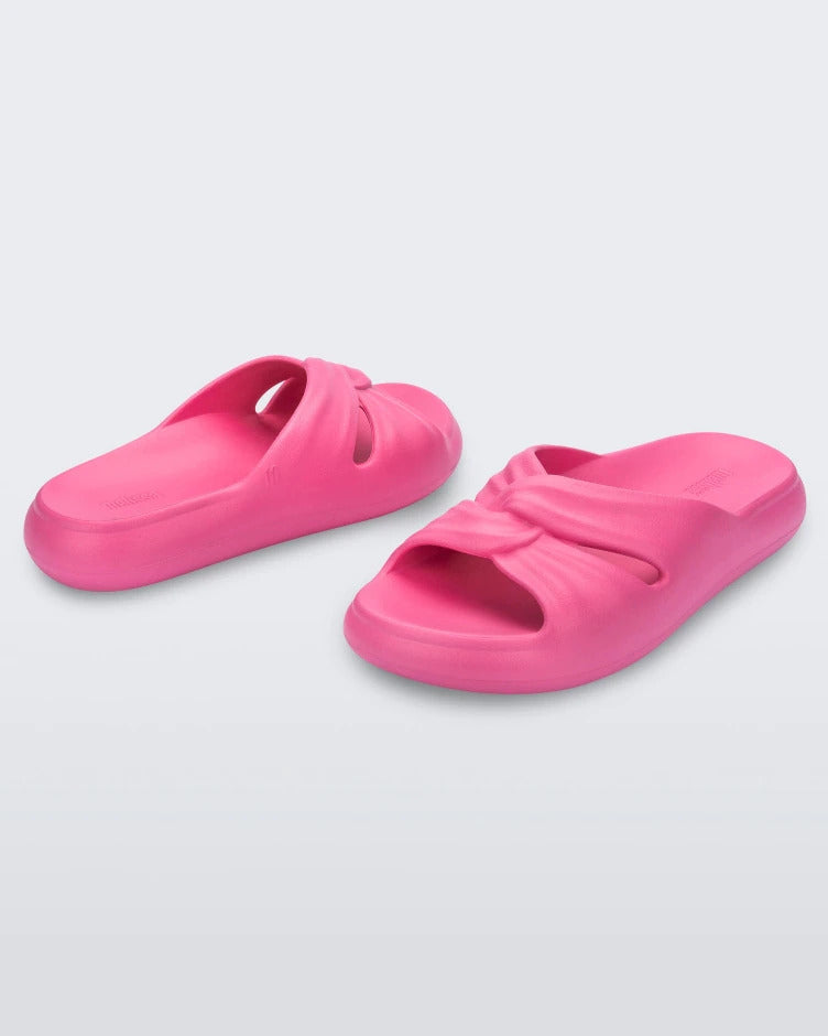An angled back and front view of a pair of pink Melissa Free Slides with a twist front detail.