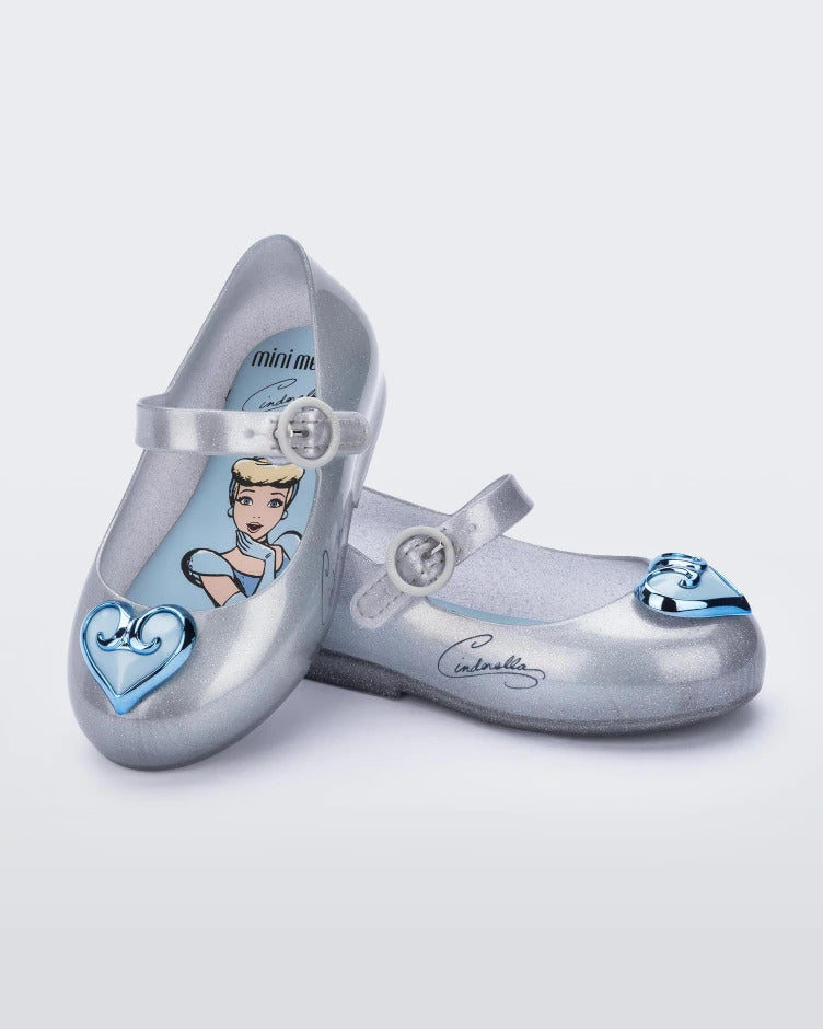An angled top and side view of a pair of silver Mini Melissa Sweet Love Princess flats with a top strap, silver base with Cinderella in script on the side, and a blue heart detail on the toe.