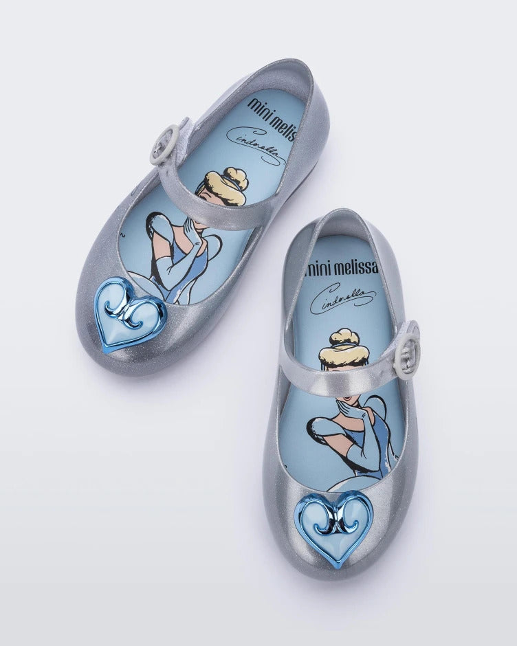 Top view of a pair of silver Mini Melissa Sweet Love Princess flats with a top strap, silver base with Cinderella in script on the side, and a blue heart detail on the toe.