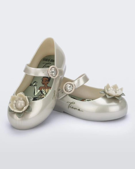 An angled top and side view of a pair of Metallic White/Green Mini Melissa Sweet Love Princess flats with a metallic white base with Tiana in script on the side, a top strap, a flower detail on the toe and a drawing of Tiana of The Princess and The Frog on the insole.