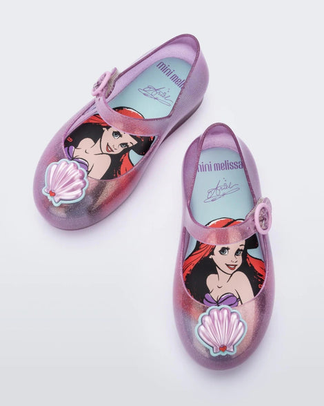 Top view of a pair of pink Mini Melissa Sweet Love Princess flats, with a pink base, a top strap, Ariel in script on the side, a shell detail on the toe, and a drawing of Ariel of The Little Mermaid on the insole.