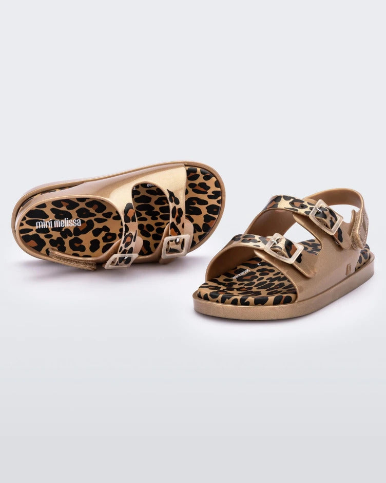 An angled side and top view of a pair of gold/brown Mini Melissa Wide Sandals with a gold base, cheetah print insole, cheetah print straps and two buckles on top.
