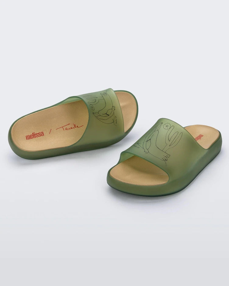 An angled front and top view of a pair of transparent green Melissa Cloud slides with a drawing of a man sitting on the ground next to a cactus and sun on the front.