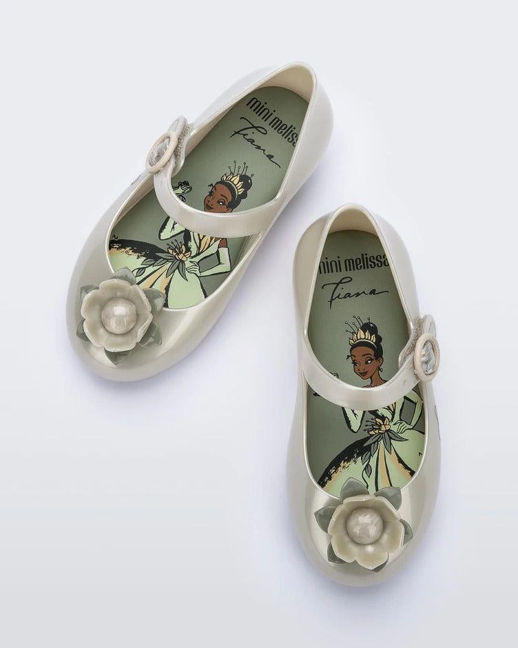 Top view of a pair of Metallic White/Green Mini Melissa Sweet Love Princess flats with a metallic white base with Tiana in script on the side, a top strap, a flower detail on the toe and a drawing of Tiana of The Princess and The Frog on the insole.