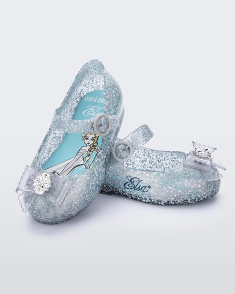 An angled top and side view of a pair of glitter blue/blue Mini Melissa Sweet Love Princess flats, with a blue glitter base, a top strap, Elsa in script on the side, a bow detail on the toe, and a drawing of Elsa of Frozen on the sole.
