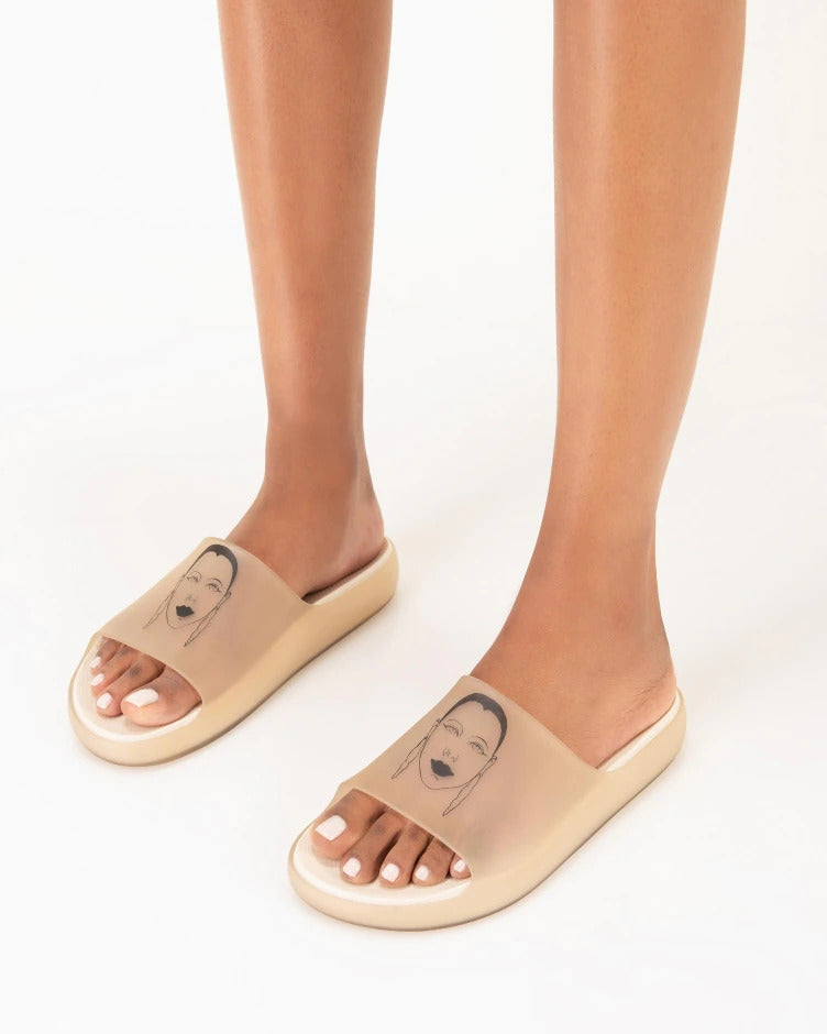A model's legs wearing a pair of transparent beige Melissa Cloud slides with a drawing of a woman's face on the front.