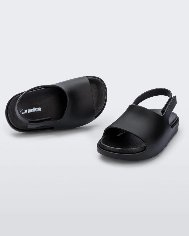 An angled top and side view of a pair of matte black Mini Melissa Cloud sandals with a black insole, base and back velcro strap.