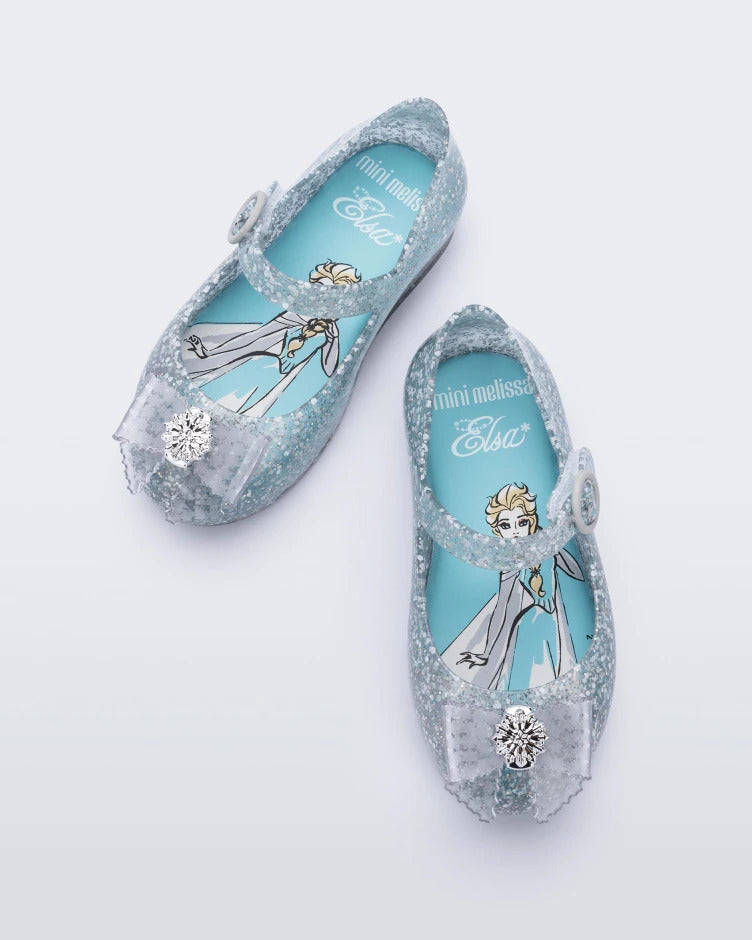 Top view of a pair of glitter blue/blue Mini Melissa Sweet Love Princess flats, with a blue glitter base, a top strap, Elsa in script on the side, a bow detail on the toe, and a drawing of Elsa of Frozen on the sole.