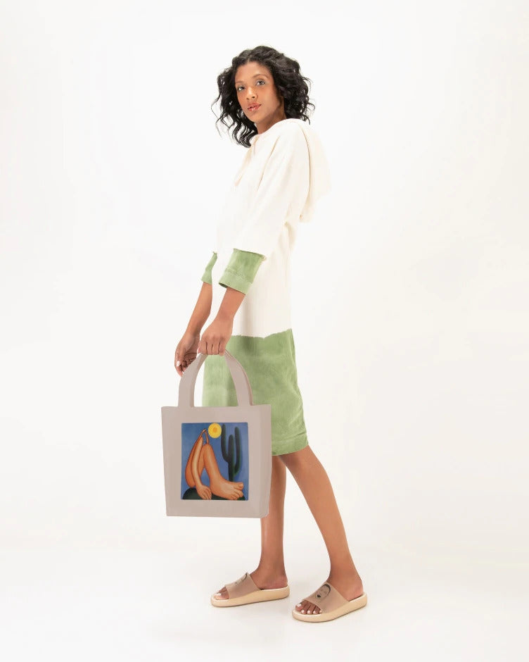 A model posing for a picture, holding a bag with a painting on it and wearing a pair of transparent beige Melissa Cloud slides with a drawing of a woman's face on the front.