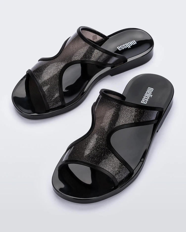 An angled top view of a pair of transparent black glitter Melissa Bikini Slides with two glitter straps conjoining in the middle.