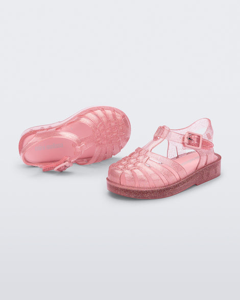 An angled top and side view of a pair of Glitter Pink Mini Melissa Possession sandals with several straps and a pink glitter base.