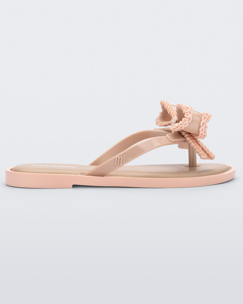Side view of a beige Mini Melissa Slim flip flop with a lace like bow detail.