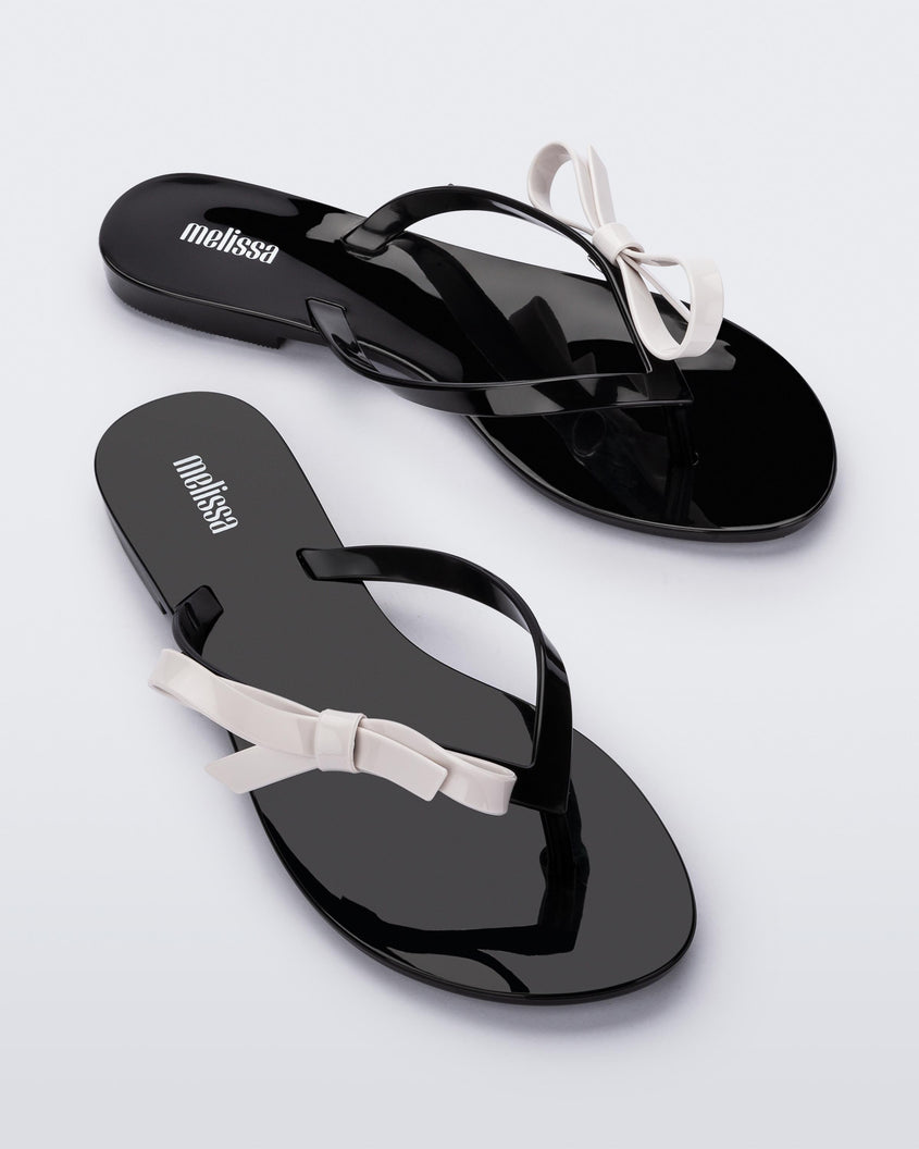 An angled top view of a pair of black/white Melissa Harmonic Sweet flip flops with a black sole, straps and white bow on top.
