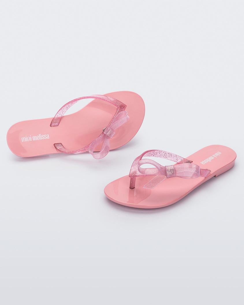 An angled side and top view of a pair of Pink/Glitter Pink Mini Melissa Harmonic Sweet flip flops with a pink sole, pink glitter straps with a bow on top.