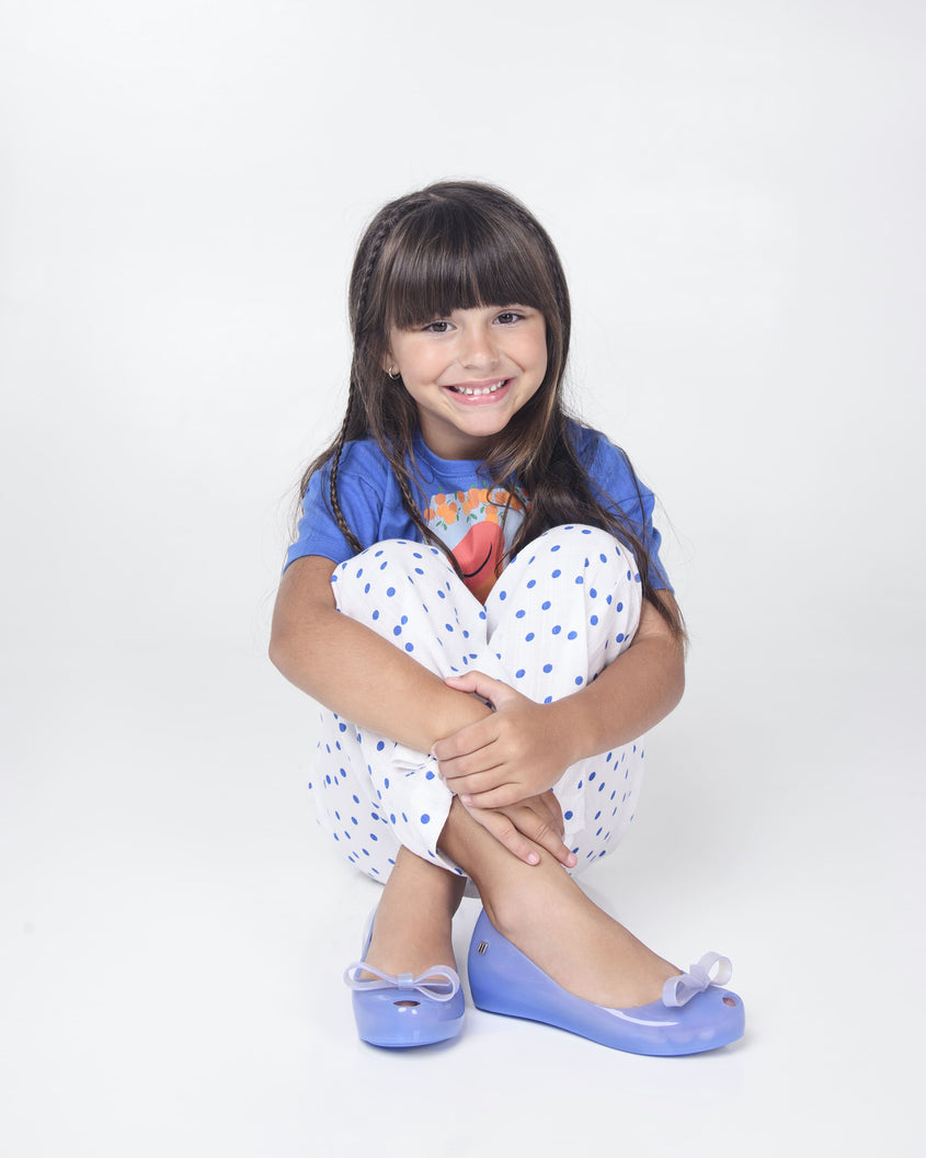 A model posing for a picture, sitting on the ground in a blue graphic top, white patterned bottoms and is wearing a pair of blue Mini Melissa Ultragirl Bow flats with a blue base and a white bow.