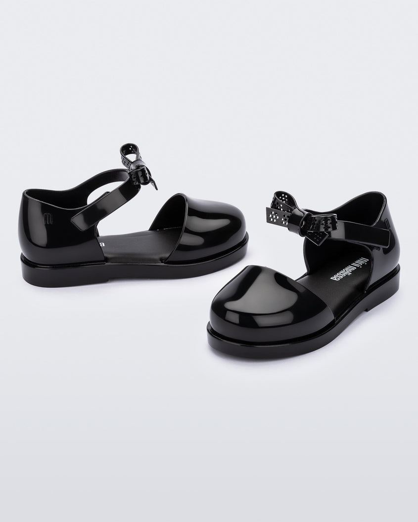 An angled front and side view of a pair of Black Mini Melissa Amy sandals with a covered toe section and a single black strap with a lace like bow detail.