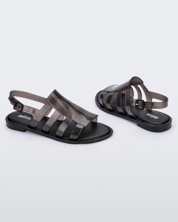An angled side and back view of a pair of transparent glitter black Melissa Boemia sandals with straps conjoining in the front and a back buckle.