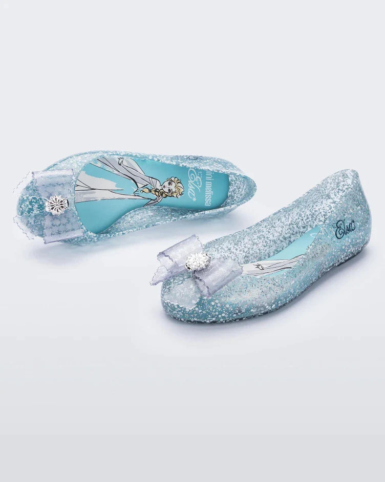 An angled front and top view of a pair of clear Mini Melissa Sweet Love Princess flats, with a clear glitter base, Elsa in script on the side, a bow detail on the toe, and a drawing of Elsa of Frozen on the sole.
