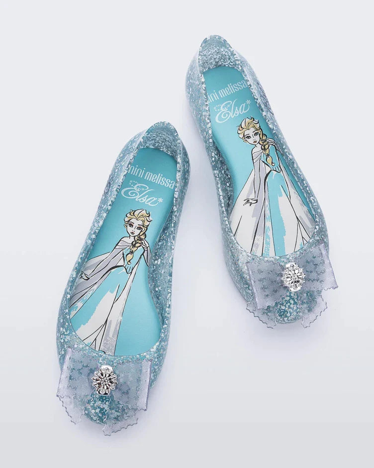 Top view of a pair of clear Mini Melissa Sweet Love Princess flats, with a clear glitter base, Elsa in script on the side, a bow detail on the toe, and a drawing of Elsa of Frozen on the sole.