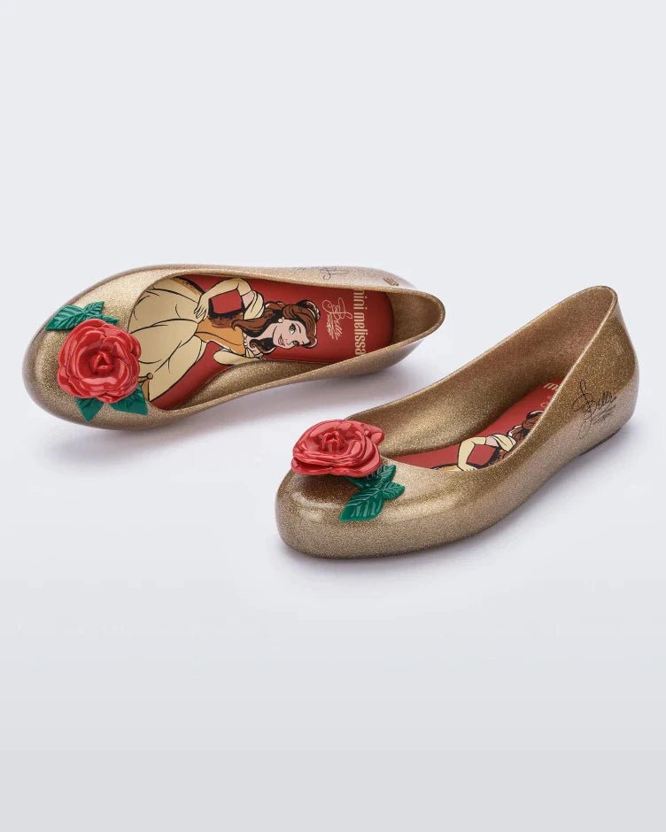 An angled front and top view of a pair of gold Mini Melissa Sweet Love Princess flats, with a gold base, Belle in script on the side, a rose detail on the toe, and a drawing of Belle of Beauty and the Beast on the sole.