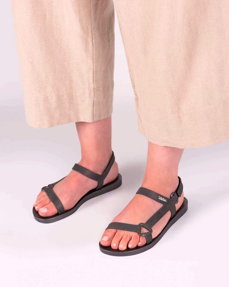 A model's legs in beige pants and a pair of graphite metallic Melissa Sun Downtown sandals with a front and ankle strap joined together by a vertical strap and a triangle buckle.