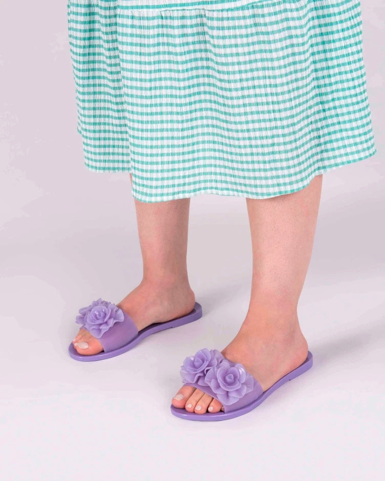 A model's legs wearing green gingham printed dress and a pair of lilac Melissa Babe Garden slides with two flowers on the front strap.