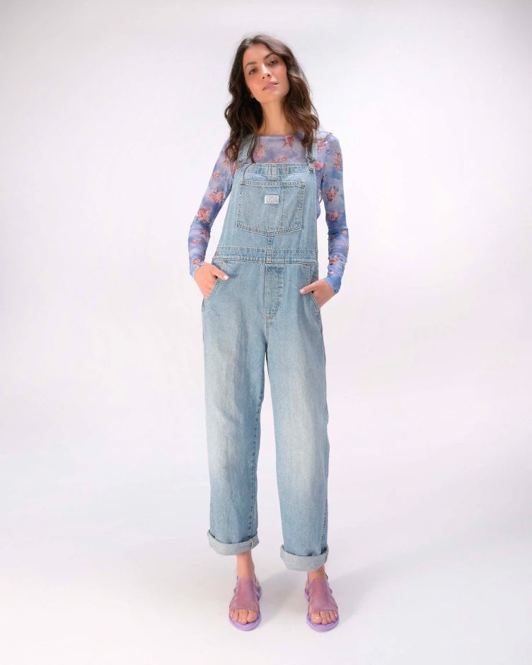A model posing for a picture in overalls and wearing a pair of transparent lilac glitter Melissa Boemia sandals with straps conjoining in the front and a back buckle.