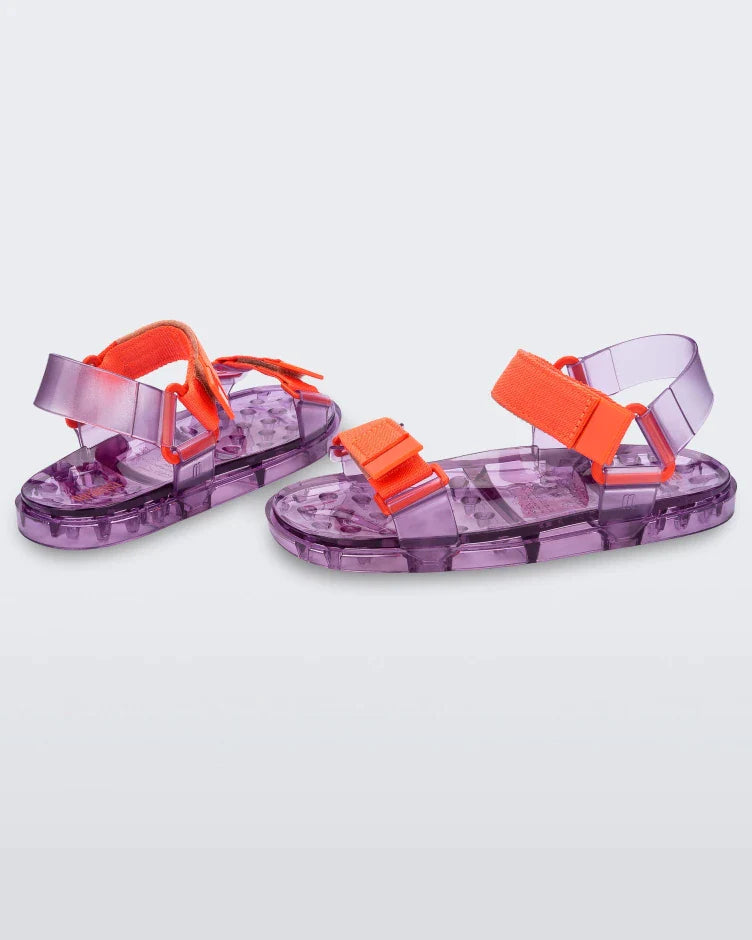 Angled view of a pair of Melissa Wide Papete sandals with transparent Purple sole and orange velcro straps. 