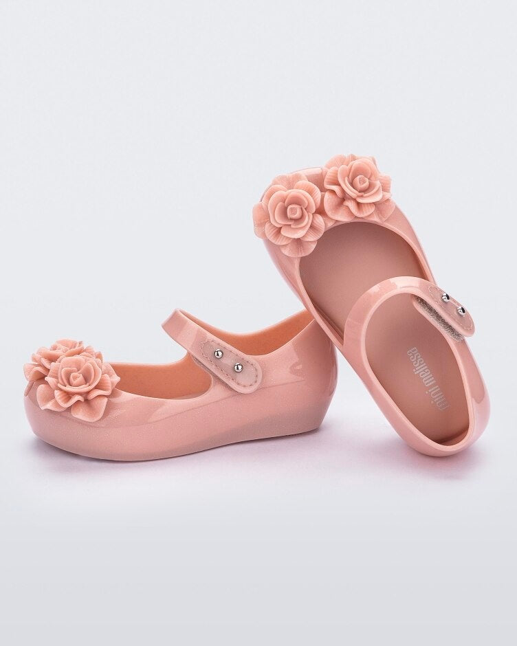 A side and top view of a pair of light pink Mini Melissa Ultragirl Garden flats with a top strap and two rose details on the toe.