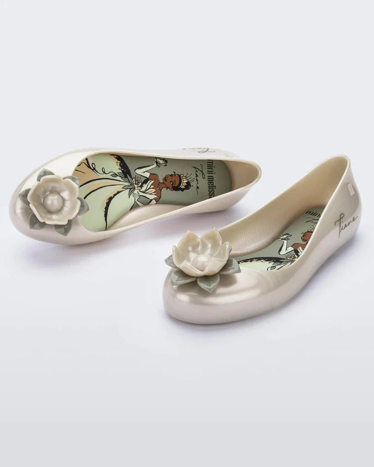 An angled front and side view of a pair of green Mini Melissa Sweet Love Princess flats, with a pearly green base, Tiana in script on the side, a flower detail on the toe, and a drawing of Tiana of The Princess and the Frog on the sole.