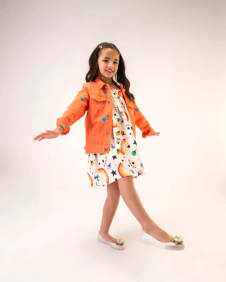 A young model in a white patterned dress, orange jacket and a pair of green Mini Melissa Sweet Love Princess flats, with a pearly green base, Tiana in script on the side, a flower detail on the toe, and a drawing of Tiana of The Princess and the Frog on the sole.