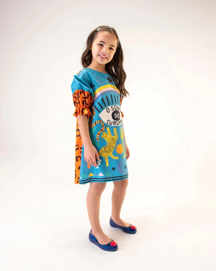 A model posing for a picture in a patterned dress and a pair of blue Mini Melissa Sweet Love Princess flats, with a blue base, Snow White in script on the side, an apple detail on the toe, and a drawing of Snow White on the sole.