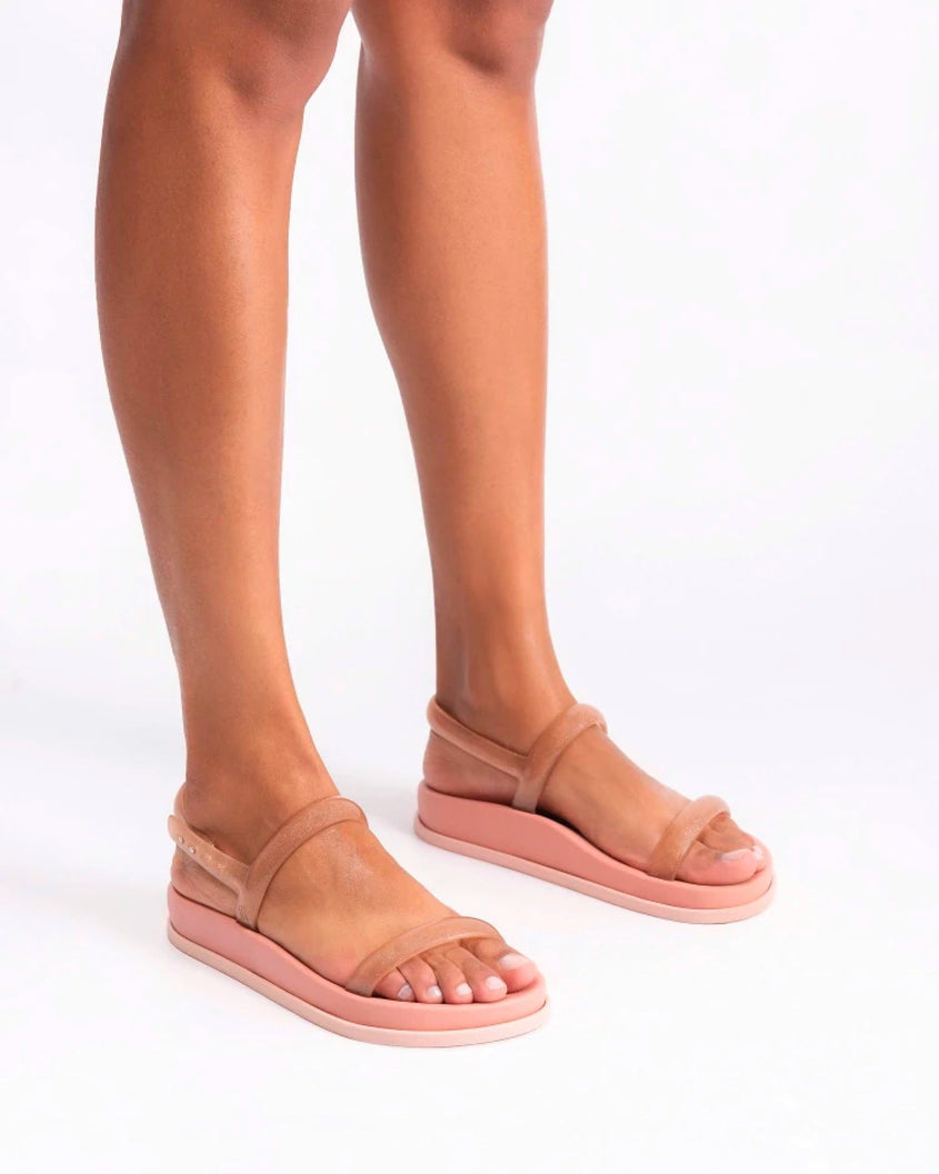 A model's legs wearing a pair of pink/clear pink Melissa Soft Wave Sandals with a pink sole, two front clear pink straps and a clear pink ankle strap.
