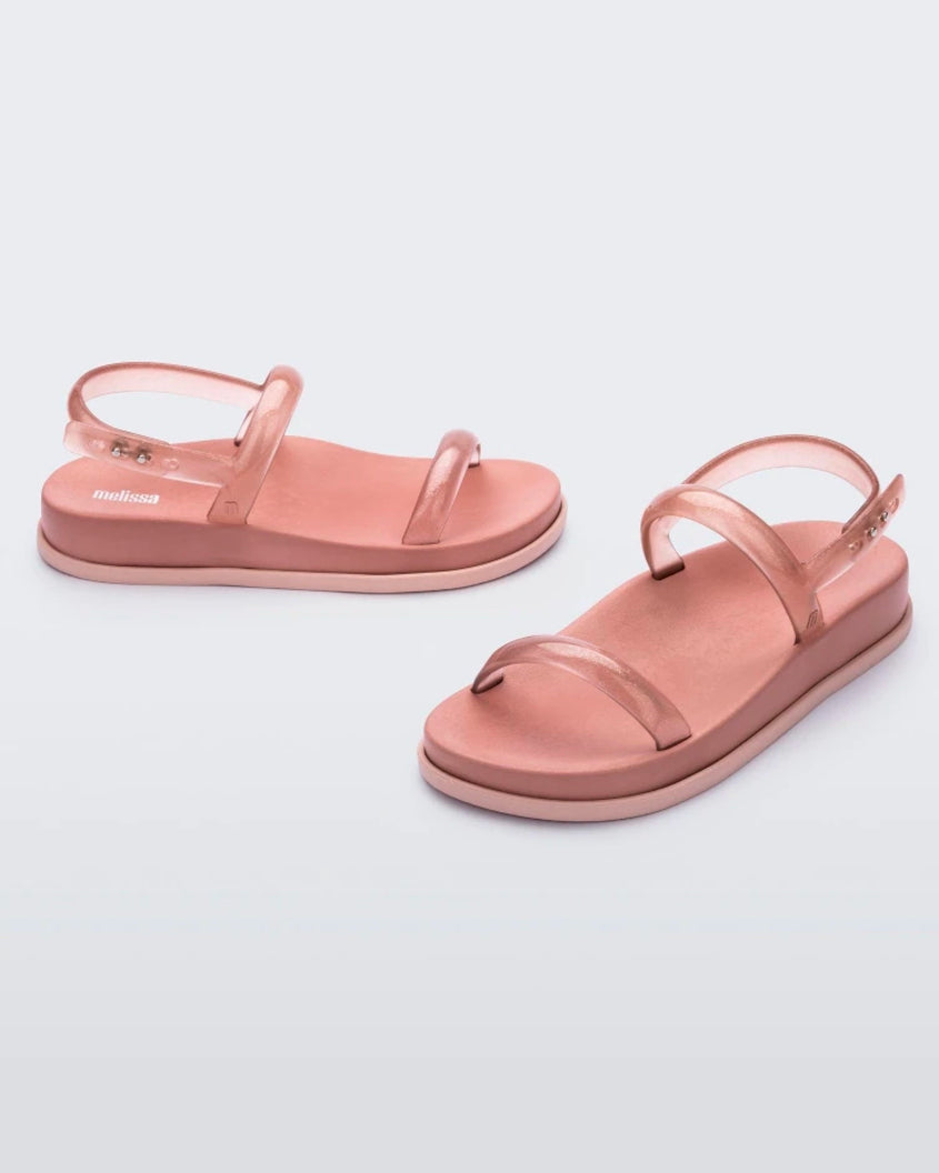 An angled front and side view of a pair of pink/clear pink Melissa Soft Wave Sandals with a pink sole, two front clear pink straps and a clear pink ankle strap.