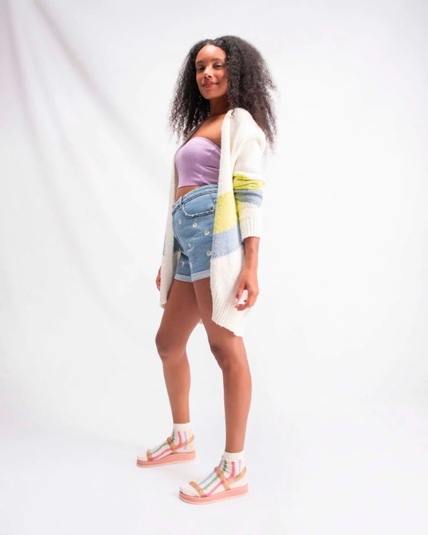 A model posing for a picture in a sweater, purple top, denim shorts and a pair of pink/clear pink Melissa Soft Wave Sandals with a pink sole, two front clear pink straps and a clear pink ankle strap.