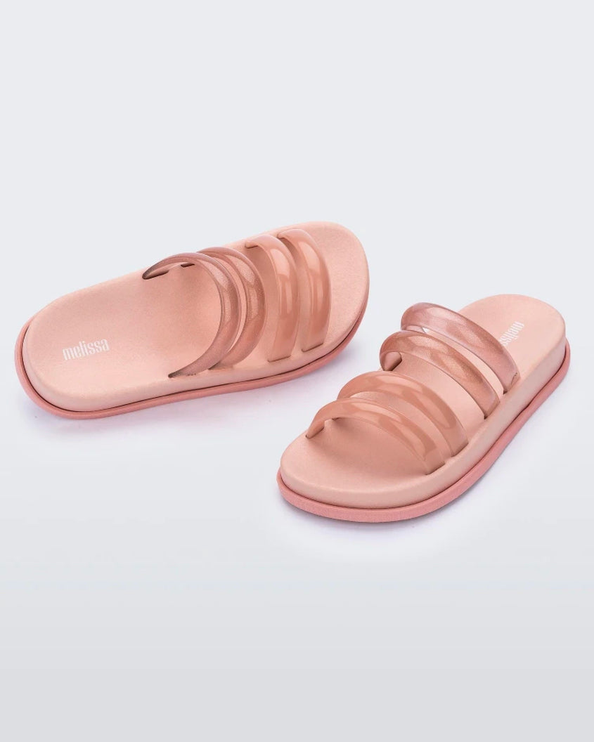 An angled front and side view of a pair of pink/clear pink Melissa Soft Wave Slides with 4 straps: two pink and two clear pink.