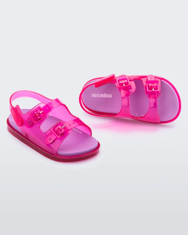 An angled front and top view of a pair of pink/lilac Mini Melissa Wide Sandals with a pink base, two pink buckles on top, a back ankle strap and a lilac insole.